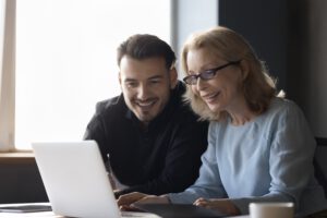 Ein Mann und eine Frau, beide Kolleg*innen, sitzen nebeneinander und blicken gut gelaunt auf einen Laptop. Die positive Stimmung zeigt, wie wertschätzende Kommunikation Zusammenarbeit stärkt und ein angenehmes Miteinander im Team fördert.