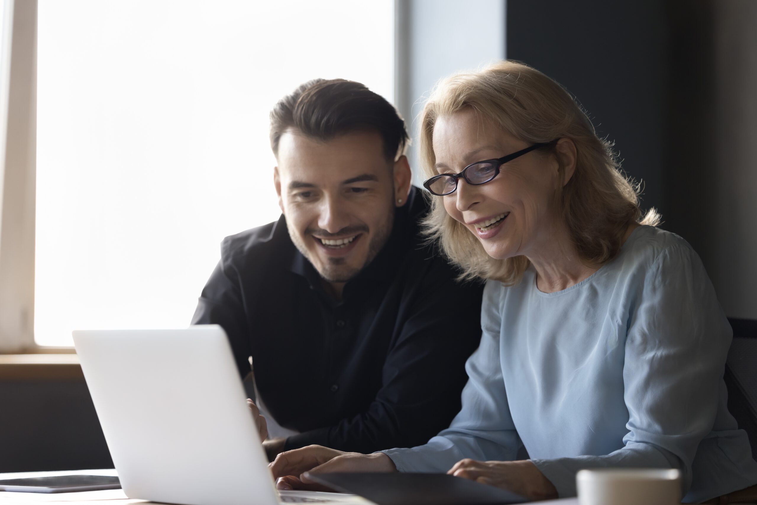Ein Mann und eine Frau, beide Kolleg*innen, sitzen nebeneinander und blicken gut gelaunt auf einen Laptop. Die positive Stimmung zeigt, wie wertschätzende Kommunikation Zusammenarbeit stärkt und ein angenehmes Miteinander im Team fördert.